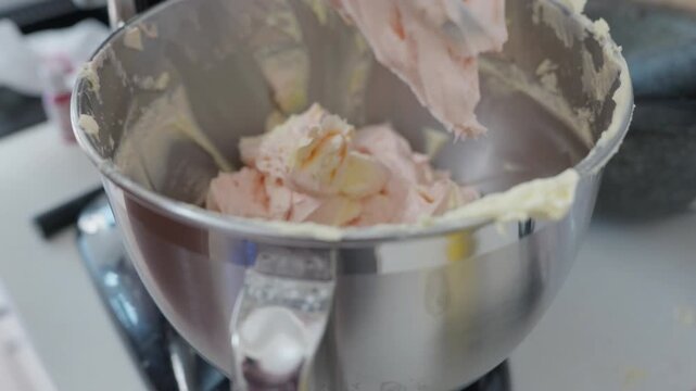 Close up, A cook taps a spatula on the side of a mixer bowl to tap off excess buttercream icing, then lowers the paddle and puts the mixer on low speed to whip the icing.