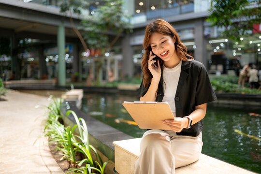 Asian businesswoman talking on phone holding clipboard in urban park