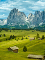 Dramatic landscape in the Dolomites, Italy. Green alpine meadows, wooden huts, and majestic mountains under a mix of sunlight and storm clouds — pure natural harmony and power.