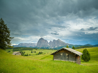 Dramatic landscape in the Dolomites, Italy. Green alpine meadows, wooden huts, and majestic mountains under a mix of sunlight and storm clouds — pure natural harmony and power.