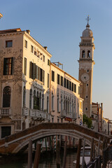 Obraz premium A charming, small brick arch bridge over a narrow Venice canal, with historic palazzo facades bathed in the warm light of the golden hour, and an ornate clock tower rising in the background