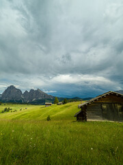 Dramatic landscape in the Dolomites, Italy. Green alpine meadows, wooden huts, and majestic mountains under a mix of sunlight and storm clouds — pure natural harmony and power.
