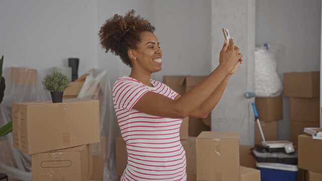 Woman holds smartphone to take selfie with raised arm in a building full of moving boxes and packed plants, smiling; moving day joy.