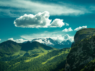 Peaceful alpine valley in the Dolomites, Italy. Rolling green hills and pine forests leading to snow-covered peaks in the distance under a soft summer sky — pure mountain serenity.