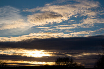 Dramatic sunset sky with vibrant golden and dark layered clouds, creating an atmospheric natural backdrop above silhouetted trees