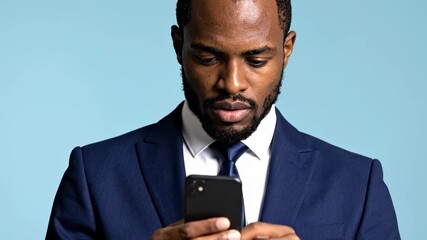 Serious young African American businessman in a formal suit checking his mobile phone for messages against a blue background - Powered by Adobe