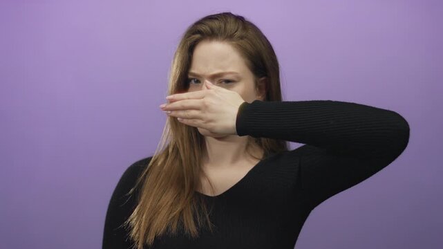 Young caucasian woman with long brown hair pinches nose and tightly closes eyes in purple studio; disgust.