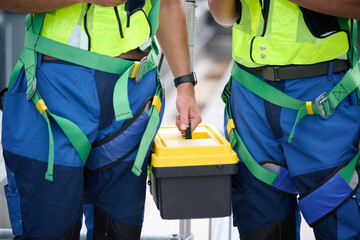 Two construction workers hands wearing safety harnesses are holding toolbox