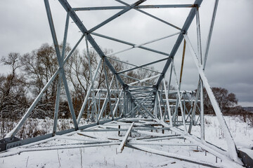 A power line pylon's metallic frame lays covered in fresh snow across a wintry field, awaiting erection. Bare trees dot the cloudy horizon