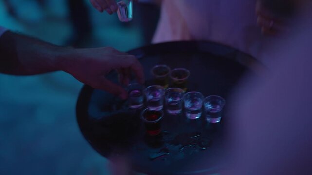 A dynamic, close-up shot captures hands taking chasers or shot glasses filled with alcohol an a dark, round serving tray. The scene is illuminated by striking neon club lights.