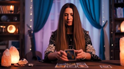 A woman with long hair, dressed in a patterned top, sits at a table, focused on tarot cards in a dimly lit room - Powered by Adobe