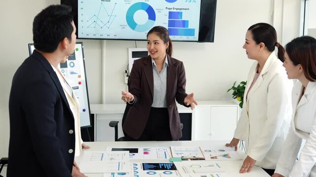 Professional businesswomen analyzing marketing data, collaborating around table with charts, graphs, and large screen displaying additional information in modern office setting