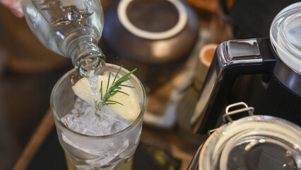 Close up of pouring fresh drink into glass with ice, lemon, and sprig of rosemary. refreshing beverage, cool cocktail being prepared in cozy cafe