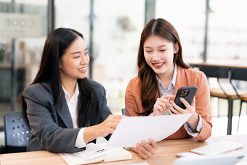 Two cheerful Asian businesswomen are collaborating, discussing documents, and reviewing information on a mobile phone during a positive meeting in a modern office.