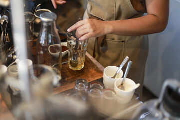 Professional barista wearing brown apron focused on making and pouring fresh tea drink into glass mug at cozy cafe counter. scene shows careful preparation
