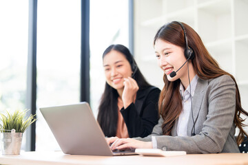 Two cheerful Asian businesswomen, one as a call center agent with a headset, are collaborating and working together on a laptop in a modern office environment.