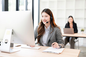 A smiling Asian female call center agent wearing a headset and working at a desktop computer, taking notes while providing customer support in a busy office environment.