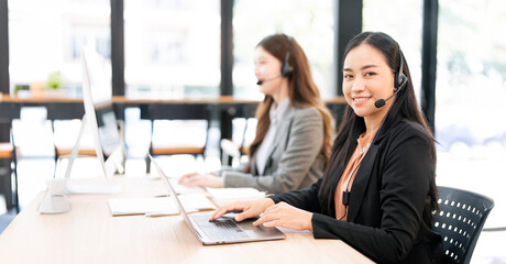 A friendly and professional Asian female call center agent wearing a headset, smiling at the camera while assisting a client on her laptop in a modern office.