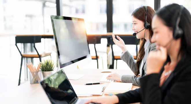 Two dedicated Asian female call center agents wearing headsets, collaborating and speaking to customers while working on computers in a bright, modern office.