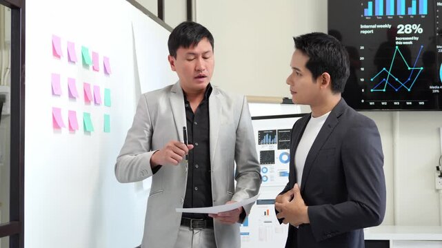 Professional businessmen collaborating in corporate meeting room, analyzing strategy with whiteboard, sticky notes, monitor displaying growth charts and performance data