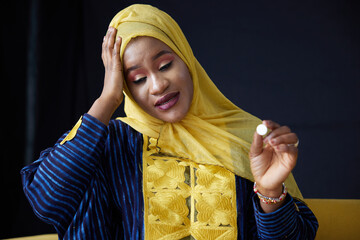 Muslim woman thoughtfully examining a single pill, wearing a yellow headscarf, with a look of...