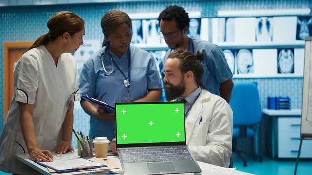 Team of doctors consulting analytics next to green screen on laptop in medical cabinet, examining x ray scan results to find the diagnosis. Licensed medics review hospital records at desk. Camera B.