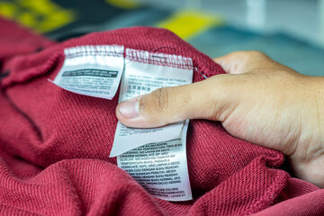 Close-up of a hand holding a red garment with visible clothing care labels, showing washing and fabric instructions in multiple languages