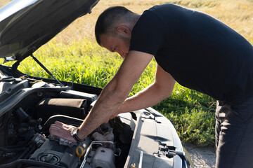 young male driver stands on street with hood open, attempting to repair it. A man checks engine of...