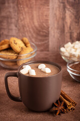 Cozy hot chocolate or sweet homemade cocoa drink decorated with marshmallows served in cup or mug on dark brown table with cinnamon and anise spices, powder ingredient and cookies as holiday dessert