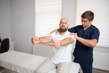 Physiotherapist guiding male patient with resistance band exercise for shoulder rehabilitation in clinic. Physical therapy session focused on injury recovery, strengthening, mobility improvement