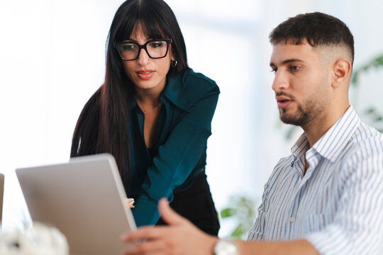 A woman wearing glasses and a man in a striped shirt collaborate on a project, looking intently at a laptop screen in a bright office setting.