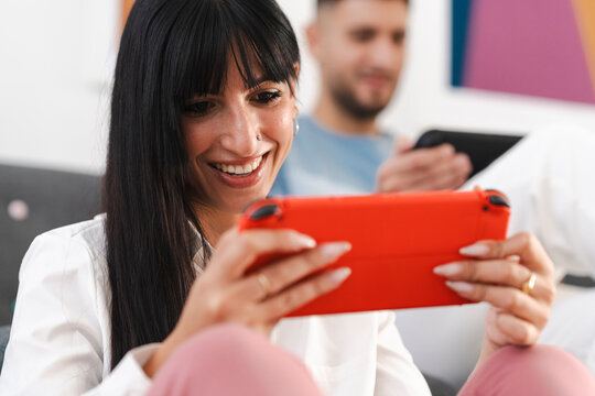 A woman smiles as she plays a video game on a handheld console, with a man in the background also engaged with a device.