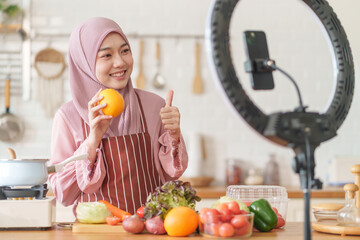 young beautiful muslim woman in hijab prepare organic fruits and vegetables for cooking on video live streaming in the kitchen,concept healthy food,technology,new marketing