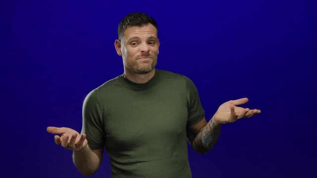 Young man with tattooed arm in green shirt shrugging and expressing confusion against blue background