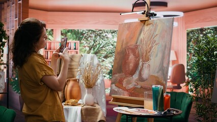 In atelier, painter is promoting her rustic masterpiece painting with the smartphone, self advertising content on social media. Filming colorful canvas with fine art piece. Online presence. Camera B.