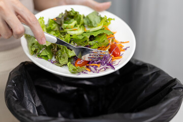 Close up of women discarding fresh vegetable salad into trash bin. Concept for food waste, diet rejection, meal dissatisfaction, eating disorder behavior or sustainability issue.