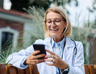 Portrait of a mature older female woman doctor or nurse and health care worker using a smartphone mobile phone in front of hospital or clinic outside