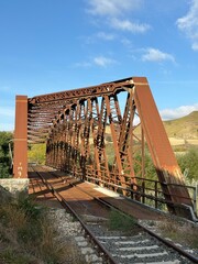An old, rusty iron railway bridge spans a valley surrounded by nature, amidst golden hills and a clear sky.