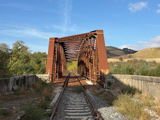An old, rusty iron railway bridge spans a valley surrounded by nature, amidst golden hills and a clear sky.
