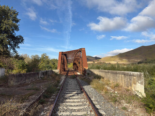 An old, rusty iron railway bridge spans a valley surrounded by nature, amidst golden hills and a clear sky.
