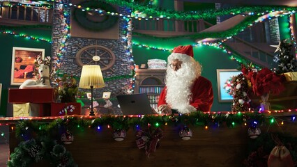 Saint Nicholas is busy online browsing Christmas websites in his festive North Pole cabin. Santa making preparations on internet webpages, spreading the magical winter holiday season. Camera B.