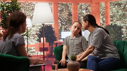 Fototapeta premium Mother bringing her zoned out dissociated teen girl at a therapy session with psychiatrist, adolescent suffering from depersonalization and disconnection from reality. Parent struggling. Camera B.
