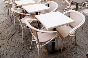 Empty white outdoor cafe tables with beige rattan chairs on cobblestone terrace pavement in European street restaurant setting