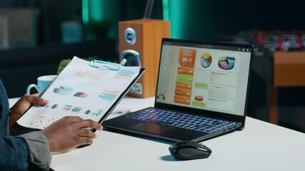 Woman sitting at home office desk, looking at business documents. African american freelancer in living room using notebook while reading financial charts on clipboard, camera A - Powered by Adobe