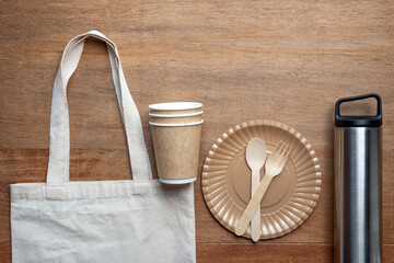 Eco-friendly flat lay with cotton tote bag, bamboo cutlery, paper cup, stainless bottle, and plate on wooden background. Perfect for zero waste, sustainable food, and takeaway concepts.