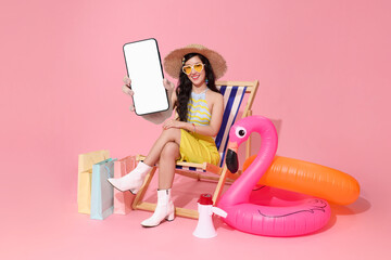 Asian woman sitting on beach chair holding smartphone blank screen and shopping bags with inflatable flamingo symbolizing summer lifestyle online shopping and digital vacation concept © NaMong Productions