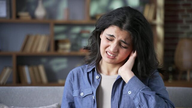 Unhealthy young woman suffers from ear pain sitting on sofa at home. Sick adult female touches her ear with fingers. She has furuncle, shingles, eczema, ear diseases or otitis media. Earache. Close up