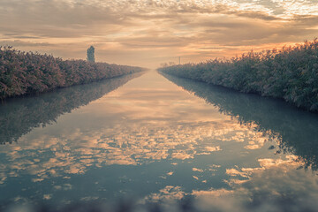 Calm rural canal at sunrise with perfect reflection of the sky and clouds on the still water. Lush vegetation lines both sides, creating a natural symmetry and peaceful atmosphere in the countryside.