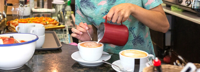 Banner of unrecognizable female barista carefully pouring steamed milk from a pitcher into a white coffee cup, creating latte art on a cappuccino in a bustling cafe setting during the day