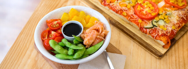 Banner of poke bowl with fresh salmon, mango, edamame, cherry tomatoes and soy sauce, serving on restaurant table next to vegetarian focaccia, in casual lunch. Organic and natural fast food concept